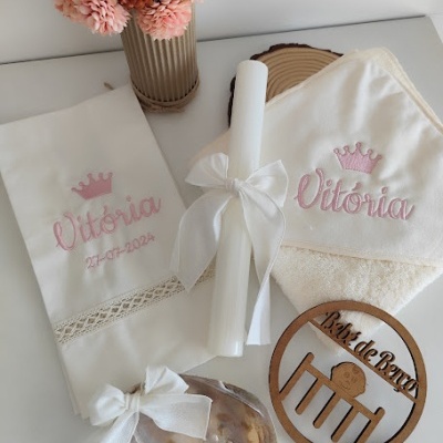 Conjunto de batismo com toalha e fralda bordadas com nome e data, vela branca com laço, sabonete e placa decorativa em fundo branco e vaso de flores.