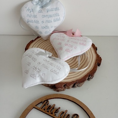 Três almofadas de tecido branco em forma de coração com bordados coloridos e texto, sobre base de madeira redonda, e ornamento de madeira com texto.