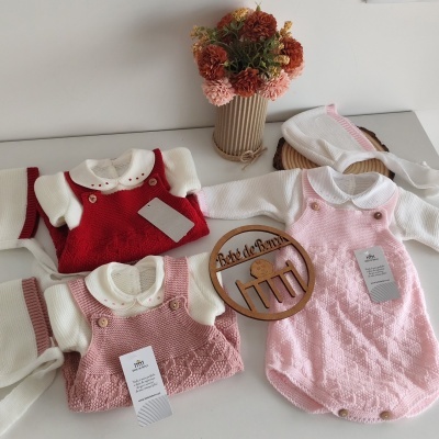 Conjuntos de roupa de bebé em malha vermelha, rosa claro e branca, sobre mesa branca com vaso de flores secas