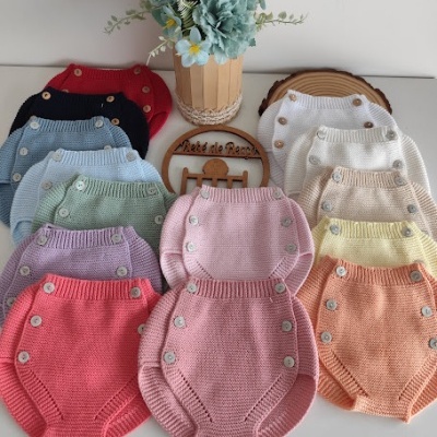 Conjunto de roupa de bebé em malha com botões laterais em várias cores sobre mesa branca com vaso de flores e placa de madeira