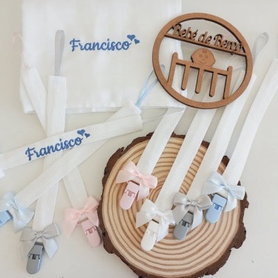 Conjunto de chupetas com fitas de tecido branco e laços coloridos, nome Francisco bordado em pano branco e placa redonda de madeira.
