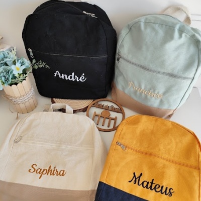Quatro mochilas coloridas com nomes bordados e um vaso de flores em fundo branco