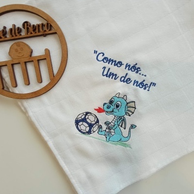 Pano de cozinha branco com dragão azul e frase, junto a objeto redondo de madeira com texto