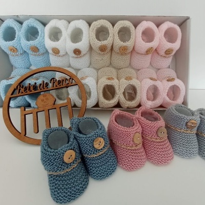 Vários pares de sapatinhos de malha para bebés com botão de madeira, organizados em caixa branca, com placa decorativa em madeira com texto Pézinhos de Lã.