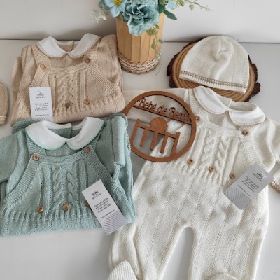 Roupas de bebé em malha bege, verde e branca com padrões trançados e boina branca ao lado de um vaso de flores azuis