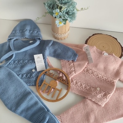 Conjuntos de roupa de bebé em malha azul e rosa com padrão de tranças, vaso com flores e enfeite de madeira