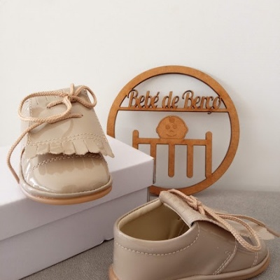 Sapatos de bebé em verniz bege com franjas e cordões sobre caixa branca, placa decorativa em madeira com texto 'Bebé de Berço' no fundo.