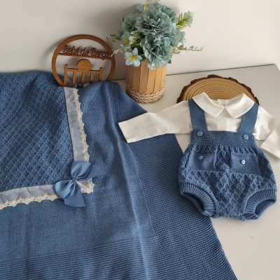 Conjunto de bebé azul com manta de tricô e roupa com botões, macaquinho azul e camisa branca, ao lado de arranjo de flores e placa de madeira