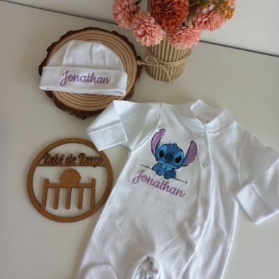 Macacão e touca brancos bordados com o nome Jonathan e personagem Stitch, sobre mesa branca com base madeira, placa redonda e vaso flores laranja e rosa