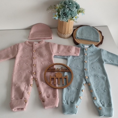 Dois macacões de bebé em malha rosa e azul claro com botões de madeira e toucas, junto a vaso de flores e decoração de madeira