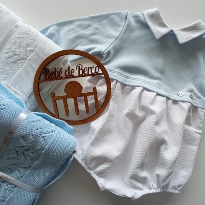 Roupa de bebé azul celeste e branca com cobertores e círculo de madeira com texto Pé de Berço
