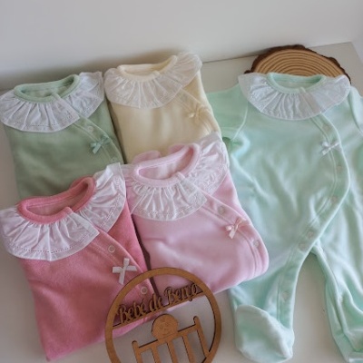 Cinco macacões para bebés em várias cores com golas rendadas brancas e laços, decorados com ornamento de madeira.