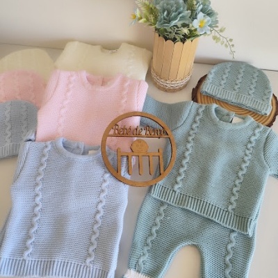 Conjunto de roupa de bebé em tricô azul, rosa e bege com padrão trançado e placa redonda de madeira com texto