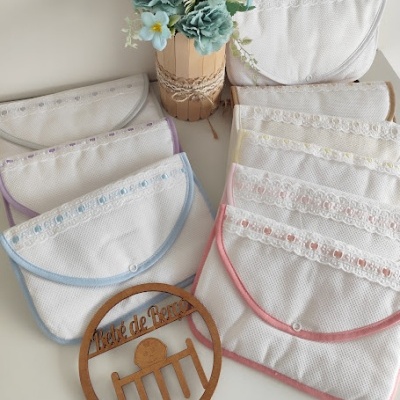 Capas de tecido branco com bordas coloridas e decoração em renda, vaso com flores azuis e círculo de madeira com texto