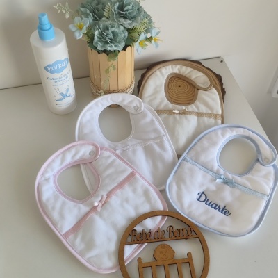 Três babetes de bebé brancos com detalhes coloridos, garrafa branca e vaso dourado com flores sobre superfície branca