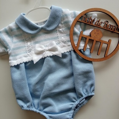 Body de bebé azul claro e branco com laço e renda, pendurado em cabide branco ao lado de placa de madeira com texto