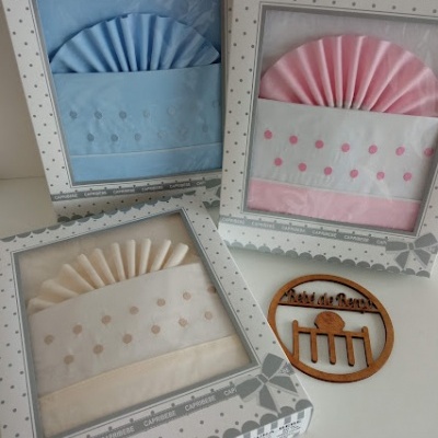 Conjuntos de roupa de cama para bebé em caixas de presente nas cores azul, rosa e creme com padrão de bolinhas bordadas, junto a ornamento de madeira redondo