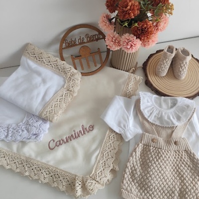 Conjunto de roupa de bebé com corpo branco e jardineiras bege de malha, manta bordada, sapatinhos e flores decorativas.