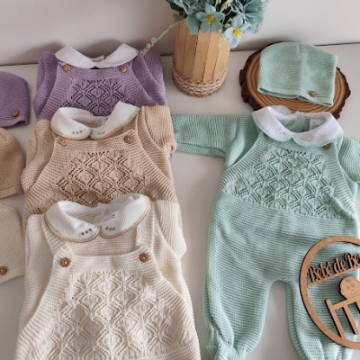 Conjunto de roupa de bebé em malha com padrões e botões, em várias cores, sobre mesa branca com flores e placa de madeira.