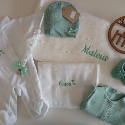 Conjunto de roupa de bebé branco e verde com nomes bordados e acessórios dispostos sobre superfície clara
