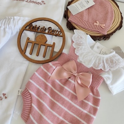 Conjunto de roupa de bebé rosa e branca com detalhes rendados e laço, com touca e meias, e fralda bordada