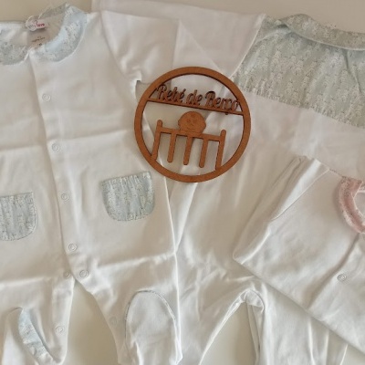 Três macacões de bebé brancos com detalhes azuis e rosas e placa de madeira com texto