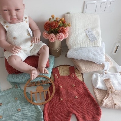 Boneca bebé com macacão de malha branco e vários macacões de malha coloridos com botões de madeira num fundo branco