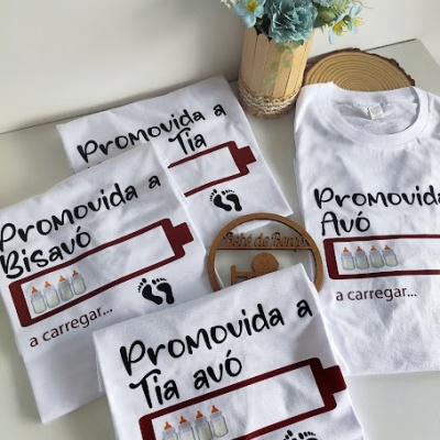 Conjunto de t-shirts brancas com texto em português e gráficos de mamadeiras e pegadas, ao lado de vaso com flores azuis.