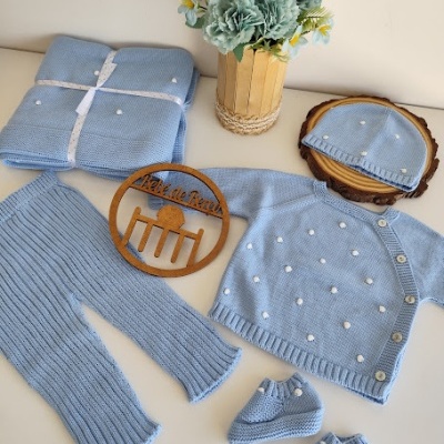 Conjunto de roupa azul claro para bebé com pompons brancos, placa de madeira e flores artificiais em vaso.