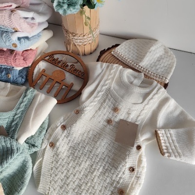 Conjunto de bebé em tricô branco com botões castanhos e gorro, roupa de bebé colorida e flores em vaso sobre superfície branca