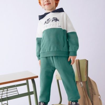 Criança vestindo conjunto verde e branco com capuz azul, desportivo cinzento, mochila bege numa cadeira verde