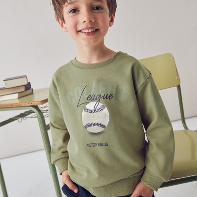 Criança com sweatshirt verde-oliva e calças azuis, junto a cadeira e secretária verdes com livros.