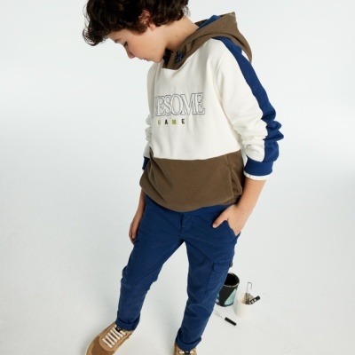 Criança com hoodie branco, castanho e azul com texto e calças azuis