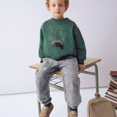 criança com camisola verde e calças cinza sentada numa mesa escolar com mochila e livros ao lado