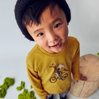 Camisola amarela com estampa de dinossauro a andar de bicicleta usada por criança com gorro preto