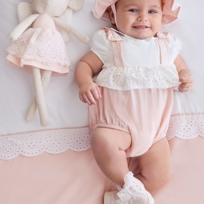Bebé vestida com fato de banho rosa claro com detalhes de renda, chapéu e sapatinhos, ao lado de boneca de pano rosa claro na cama.