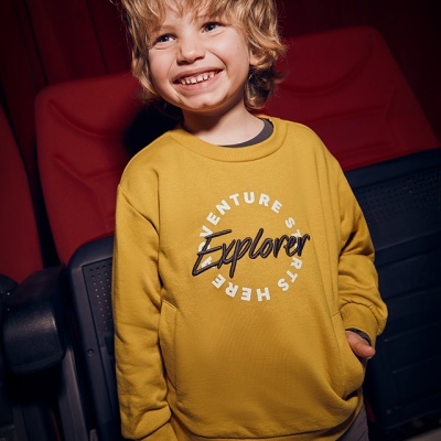 Camisola amarela com texto 'Adventure starts here Explorer' usada por criança sorridente