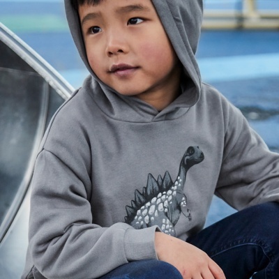 Menino com casaco cinzento com estampa de dinossauro e calças azuis num parque infantil