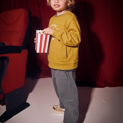 Criança a segurar caixa de pipocas em cinema com camisola amarela, calças cinza e ténis beige
