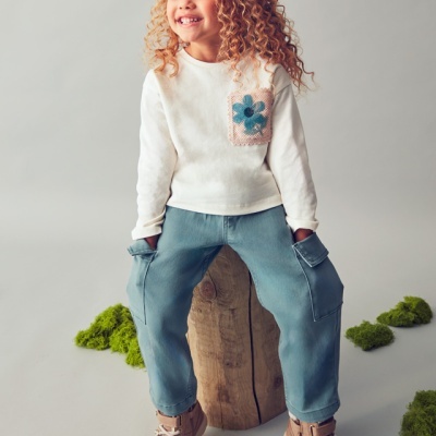 Criança com camisola branca, calças azuis e sapatilhas rosa, sentada num tronco de madeira