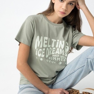 Jovem com t-shirt verde e calças de ganga sentada no chão em fundo branco.