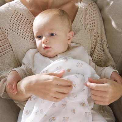 Bebé com manta estampada num sofá cinzento, com mulher de camisola bege.