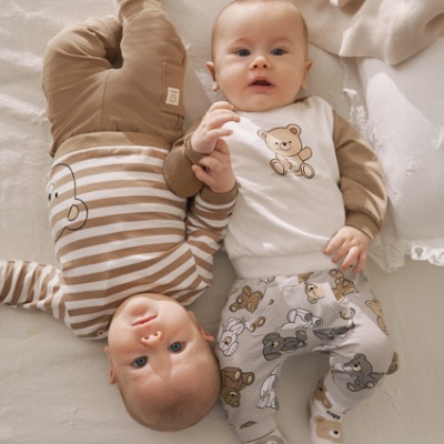 Dois bebés vestidos em tons bege e castanho deitados numa cama clara