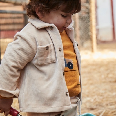 Criança vestida com roupa bege e boina azul a segurar uma pá azul num ambiente rural