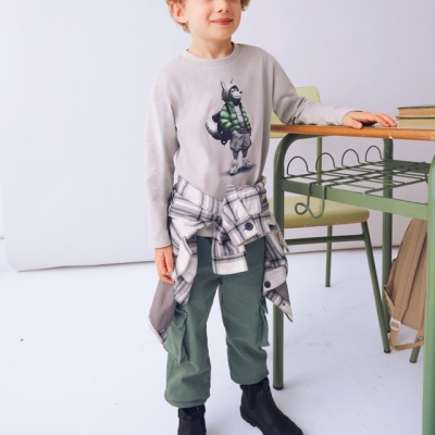 Criança em roupa casual com t-shirt cinzenta e estampa, calças verdes e botas pretas em ambiente interior