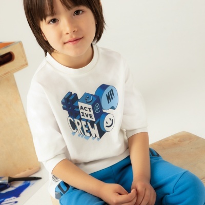 Criança com camisola branca estampada e calças azuis sentada em madeira clara.