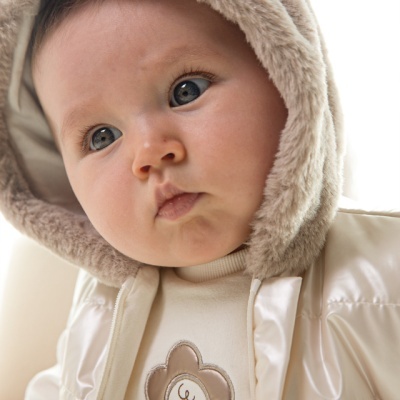 Bebé com casaco acolchoado bege e capuz de pelo sintético castanho claro e camisola creme com estampa de flor sorridente.