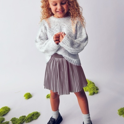 Criança com camisola cinza, saia plissada castanha, meias brancas e sapatos pretos em fundo branco com musgo verde.