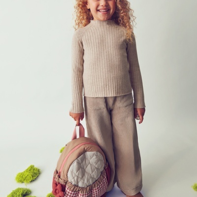 Menina com camisola e calças bege, mochila rosa e bege com padrões, fundo branco com musgo verde