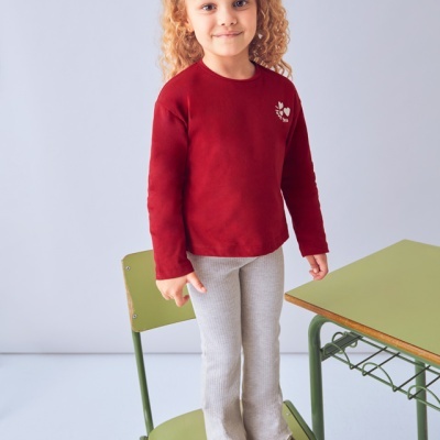 Criança com camisola vermelha, calças cinza e sapatilhas em cadeira verde ao lado de mesa verde